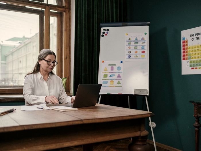 Kobieta w średnim wieku pracuje przy laptopie w sali warsztatowej, obok flipchart z materiałami.
